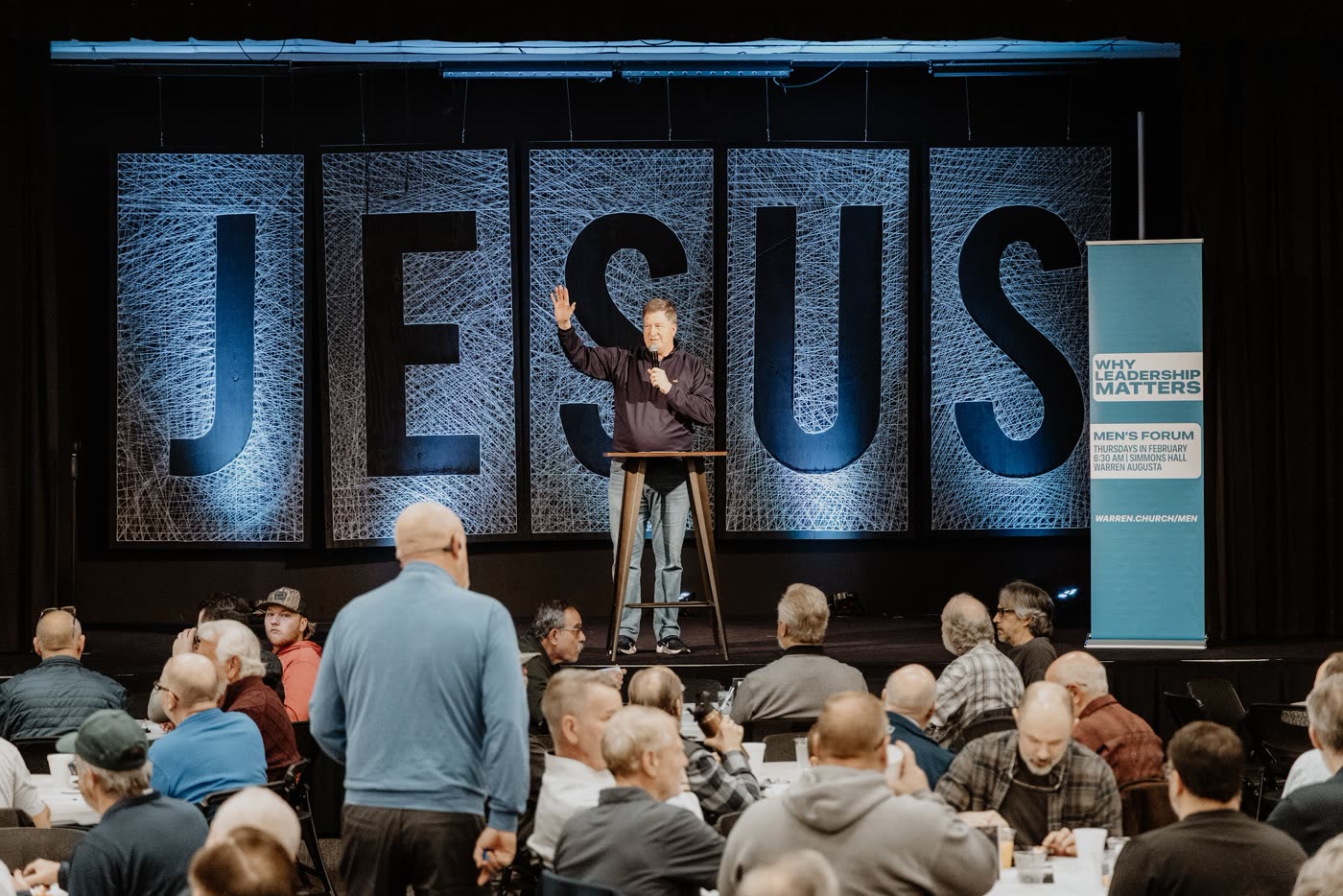 Men gathered at tables for a forum beneath large JESUS letters at Warren